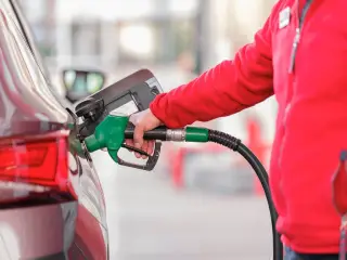 Illustration: A person in a red jacket refuels a car with a green fuel nozzle at a gas station.