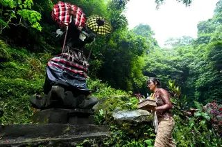 Illustration: A Balinese woman in traditional attire carrying offerings near an outdoor shrine surrounded by greenery.