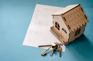 Illustration: A miniature wooden model house, keys, and a document on a blue surface.