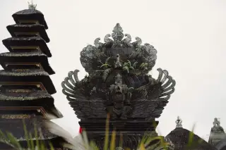 Illustration: Close-up of an ornate Balinese stone carving against an overcast sky beside a multi-tiered meru tower.