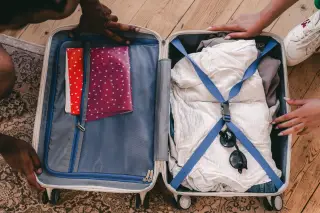 Illustration: Top-down view of an open suitcase packed with clothes and sunglasses held by two people.
