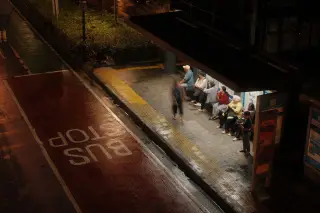 Illustration: People waiting at an illuminated bus shelter along an empty road at night.