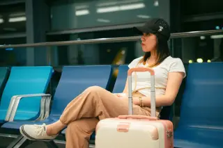Illustration: A woman wearing a cap sits on blue airport seats next to a small suitcase.