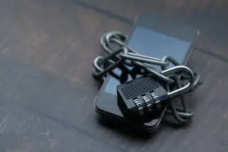 Illustration: A smartphone bound with a metal chain and locked with a combination padlock on a wooden surface.