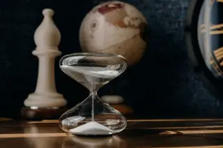 Illustration: A clear glass hourglass running with sand on a wooden surface next to a chess piece and a globe.