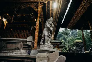 Illustration: A stone guardian statue stands beside an ornate traditional Balinese temple pavilion.