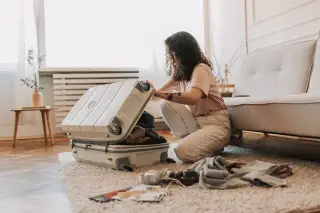 Illustration: A woman kneels on the floor packing clothes into an open suitcase inside a bright room.