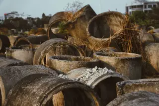 Illustration: Large weathered concrete drainage rings piled outdoors with overgrown dry weeds.
