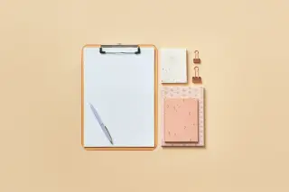 Illustration: A flat lay of a clipboard, pen, notebooks, and binder clips on a beige background.