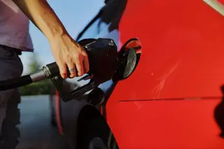 Illustration: A close-up shot of a person inserting a black fuel nozzle into a red car.