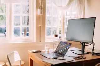 Illustration: A wooden desk with a laptop, monitor, and notebook by a bright sunlit window.