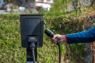 Illustration: A person's hand holding an electric vehicle charging cable plugged into a charging station outdoors.
