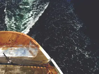 Illustration: The bow or deck of a boat moving through dark sea water with foaming wake.