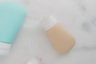 Illustration: Pastel squeezable travel toiletry bottles lying on a light marble surface.