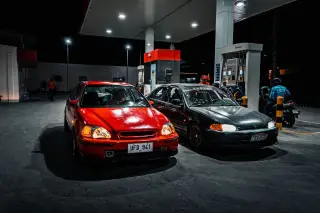 Illustration: Two cars parked side by side at a brightly lit petrol station pump at night.