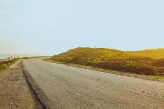 Illustration: An empty asphalt road curves beside green coastal hills under bright daylight.