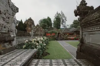 Illustration: A Balinese temple courtyard featuring stone structures, manicured lawn, and flower borders under an overcast sky.