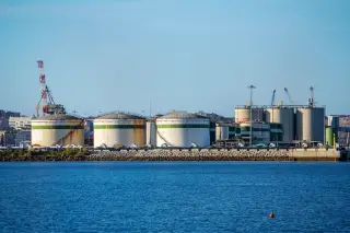 Illustration: Large industrial storage tanks and cranes at a coastal port facility viewed across blue water.