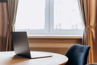 Illustration: A laptop sits on a round wooden table by a bright window with curtains.