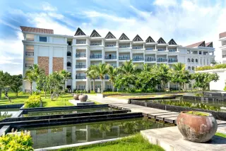 Illustration: A modern white resort building with tiered ponds and palm trees under a bright blue sky.