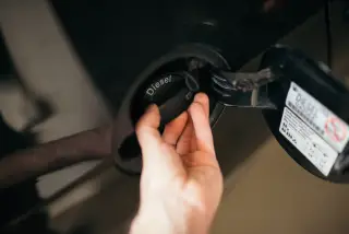 Illustration: A close-up of a person's hand opening the diesel fuel cap of a car.