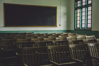 Illustration: An empty classroom with rows of wooden chairs, a blackboard, and windows.