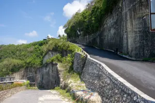 Illustration: A newly paved road climbs along a carved limestone cliff under a bright blue sky.