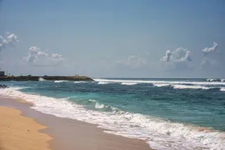 Illustration: Waves roll onto a sandy beach under a bright sunny sky with distant figures on a breakwater.