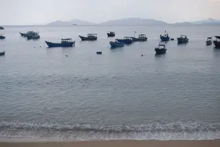 Illustration: Several wooden fishing boats anchored in calm sea waters near a sandy shore under a hazy sky.
