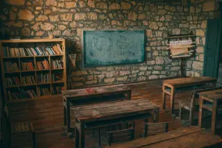 Illustration: An empty rustic classroom with wooden desks, a bookcase, and a stone wall.