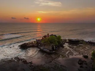 Illustration: Aerial sunset view over Tanah Lot temple on a rocky outcrop surrounded by ocean waves.