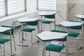 Illustration: Empty modern school desks with green chairs, exam papers, and pencils in a classroom.