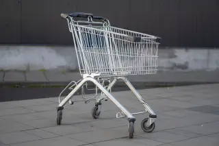 Illustration: An empty metal shopping cart stands outdoors on a paved area against a dark wall.
