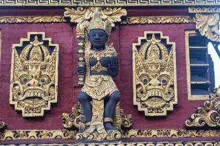 Illustration: Intricate Balinese stone carving of a deity and mythical reliefs on a red wall with gold details.