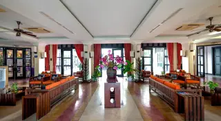 Illustration: A spacious hotel lobby with wooden sofas, orange cushions, and a large floral arrangement in the center.