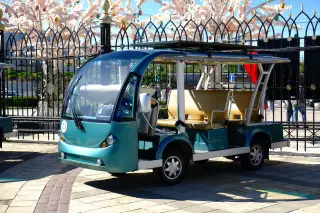Illustration: A green open-sided electric shuttle vehicle parked outdoors near an ornate fence.
