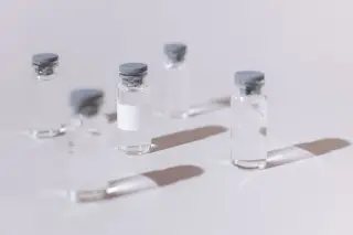 Illustration: Clear glass vaccine vials with rubber stoppers cast shadows on a plain white surface.