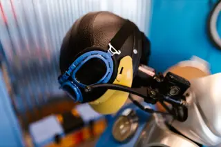 Illustration: A vintage motorcycle helmet with blue goggles resting on a scooter handlebar indoors.