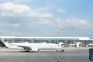 Illustration: A white passenger airplane parked on an airport tarmac in front of a glass terminal.