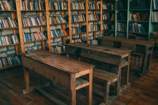 Illustration: Empty rustic wooden school desks and benches in front of tall bookshelves filled with books.