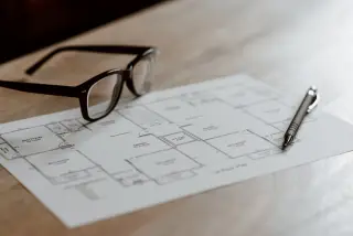 Illustration: Eyeglasses and a pen resting on an architectural floor plan on a wooden desk.
