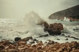 Illustration: Rough sea waves crash heavily against rocky formations along a coastal shore.
