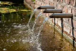 Illustration: Clear water pours from three metal pipes on a brick wall into a pond.