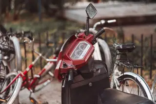 Illustration: Close-up view of the handlebar and dashboard of an old red scooter parked near bicycles.