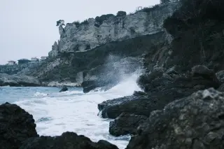 Illustration: Rough sea waves crashing against dark rocks beneath a steep cliff.