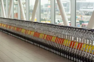 Illustration: A long neat row of luggage trolleys inside an airport terminal by large windows.