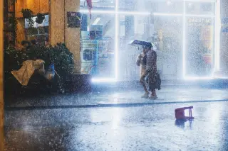 Illustration: Two people under an umbrella walk through torrential rain past brightly lit shopfronts at night.