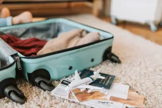 Illustration: An open turquoise suitcase packed with clothes next to a toy airplane, passport, and travel tickets.