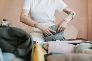 Illustration: A person with tattooed arms packing clothes into a suitcase indoors.