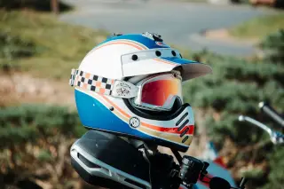 Illustration: A colorful motocross helmet and goggles resting on a motorcycle outdoors.